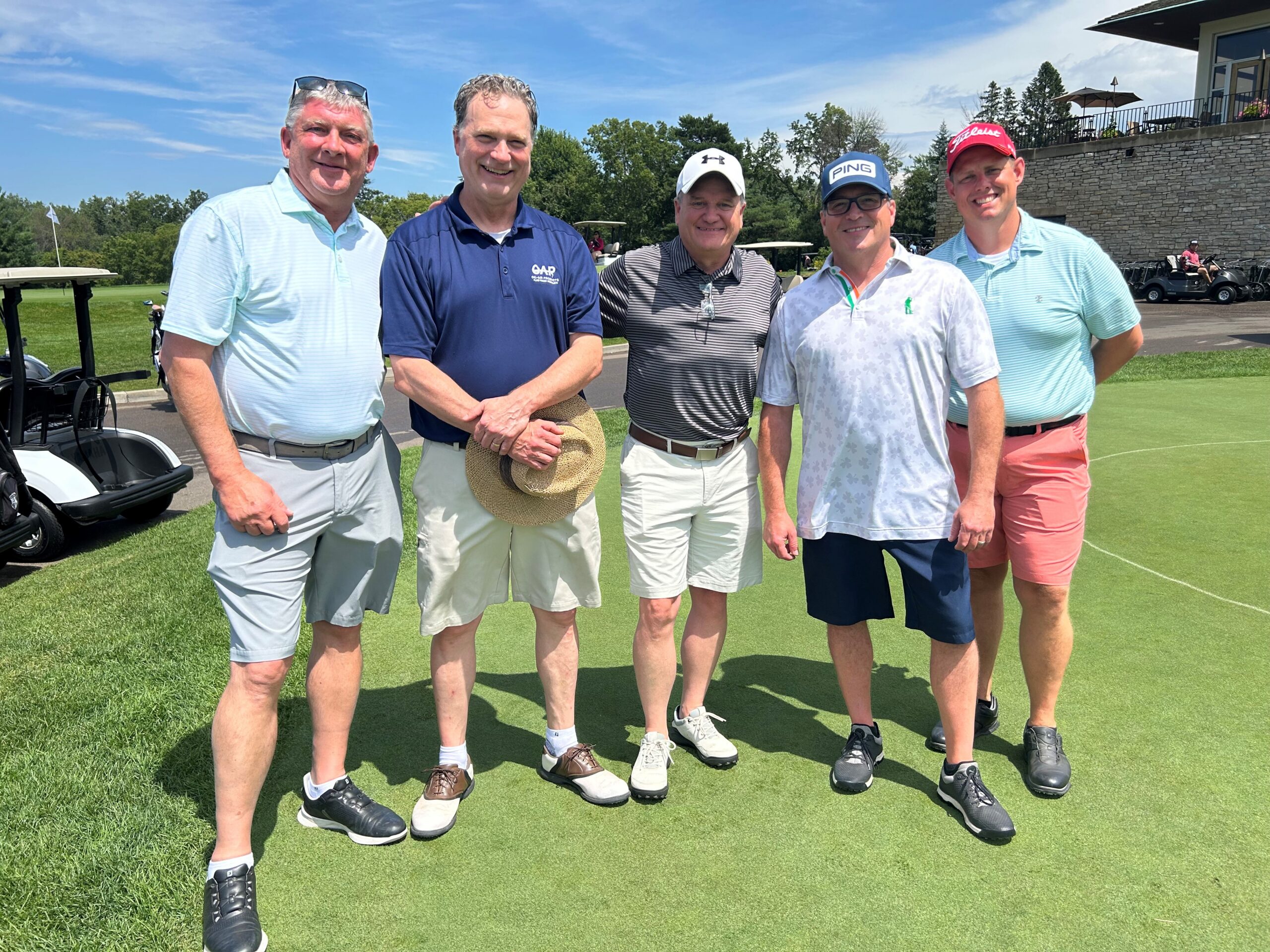 Five men in golf attire stand smiling on a green, with a golf card in the background