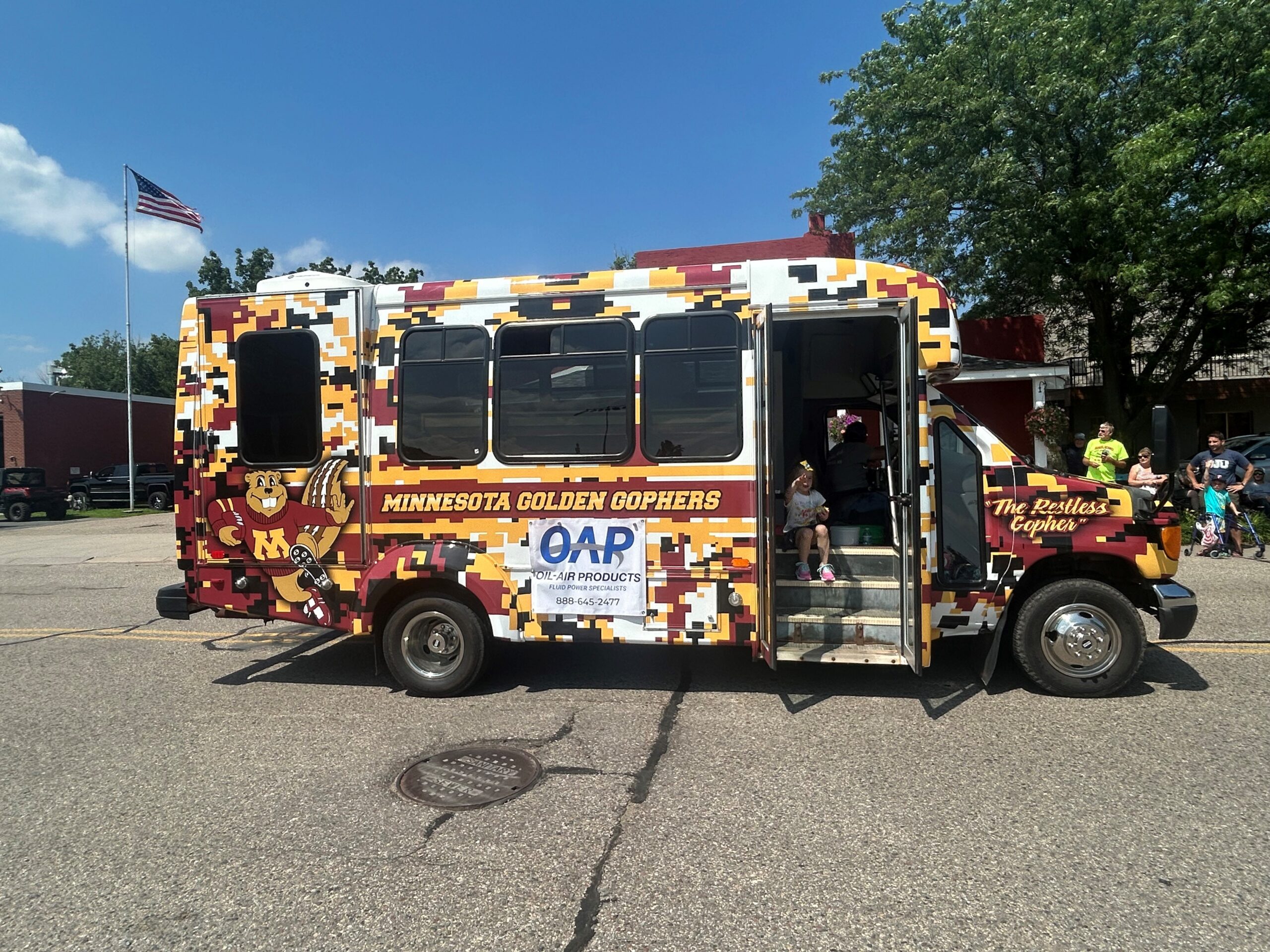 A Minnesota Golden Gophers branded mini-bus labeled "The Restless Gopher" with an Oil Air Products banner on the side and a child sitting on the bus steps